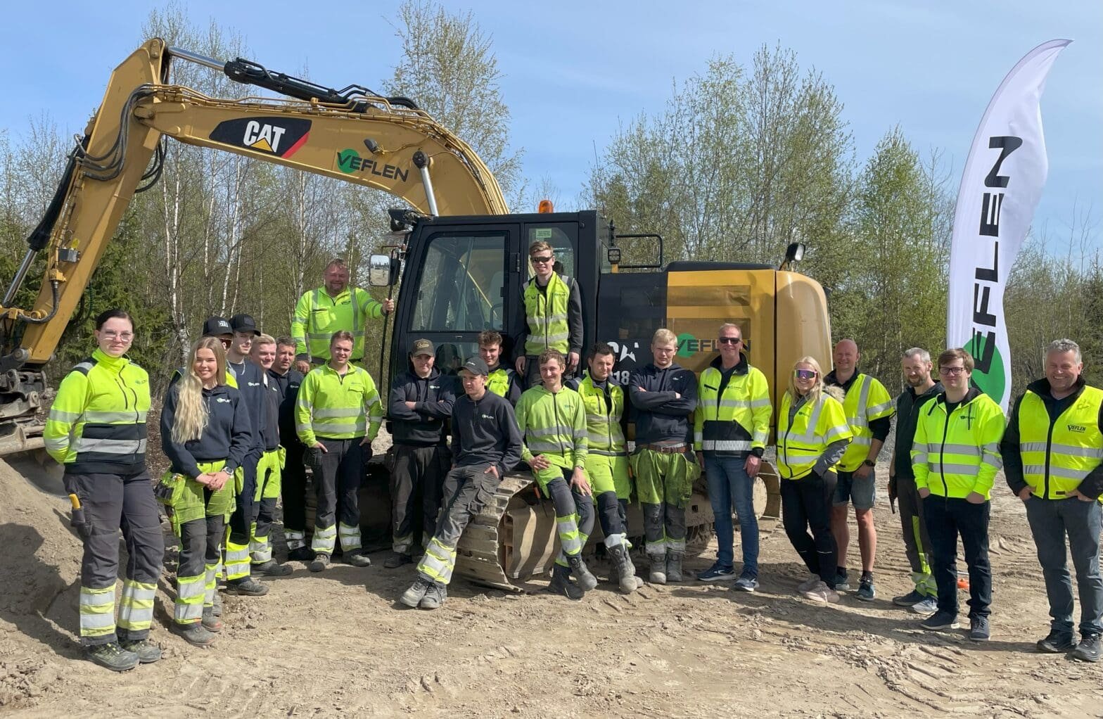 Gruppebilde av lærlinger, mentorer og ledere foran anleggsmaskiner, tatt under en vellykket lærlingedag i vårsola hos Veflen.